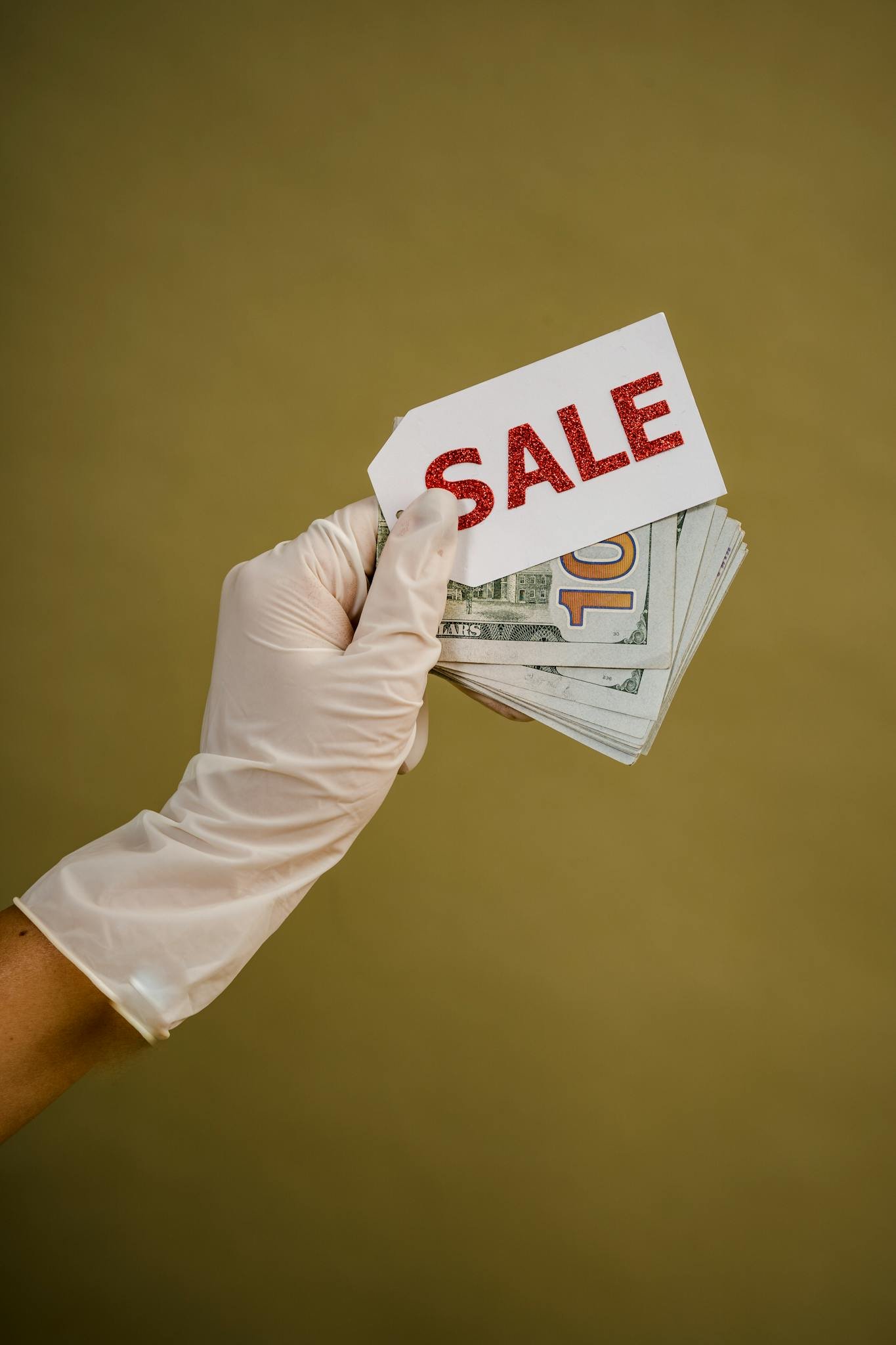 A gloved hand holds a stack of US dollars with a red 'SALE' tag against a plain background.